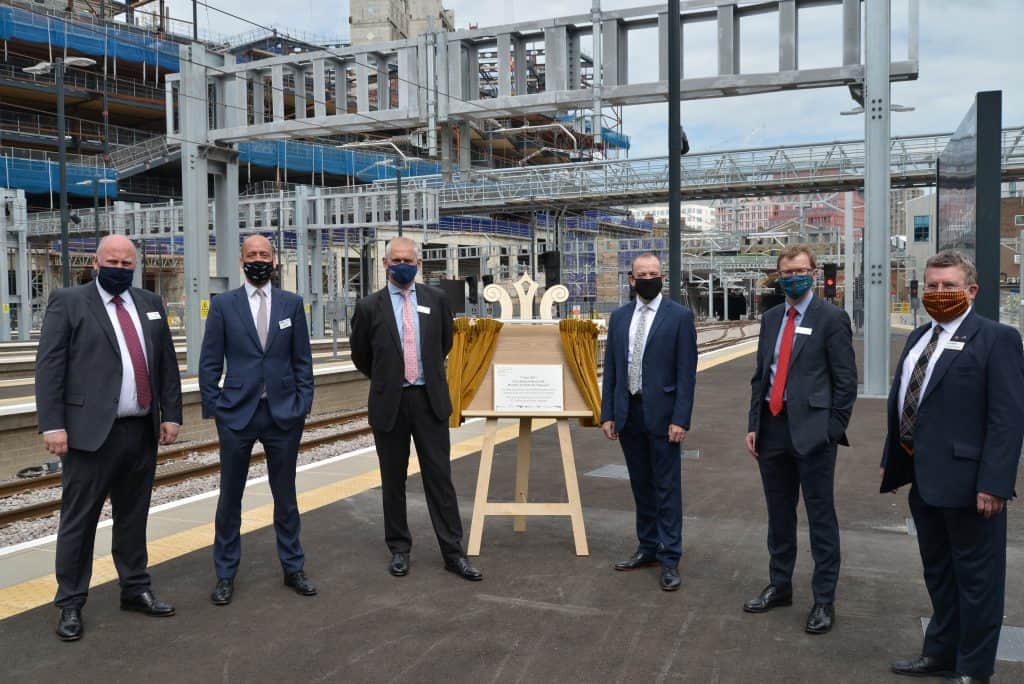 Five men in suits and face masks stnad around plaque at railway platform